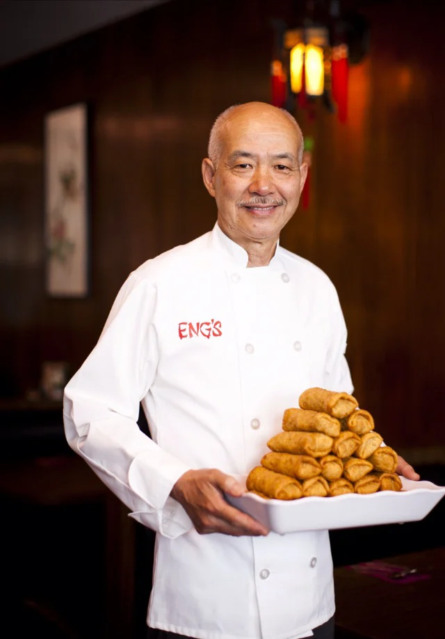 A chef in whites with ENG’S embroidered in red on the chest, holding a platter of egg rolls under a lantern.